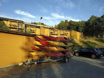Several colorful kayaks are stacked on a trailer parked beside a yellow wooden building. Apartment buildings with matching yellow exteriors are seen in the background, surrounded by trees. A black car is also parked nearby on the gravel surface. The scene is set under a clear blue sky with a few clouds.