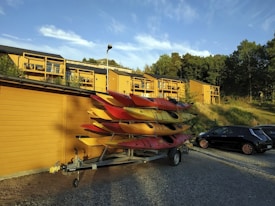 Several colorful kayaks are stacked on a trailer parked beside a yellow wooden building. Apartment buildings with matching yellow exteriors are seen in the background, surrounded by trees. A black car is also parked nearby on the gravel surface. The scene is set under a clear blue sky with a few clouds.