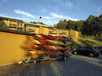 Several colorful kayaks are stacked on a trailer parked beside a yellow wooden building. Apartment buildings with matching yellow exteriors are seen in the background, surrounded by trees. A black car is also parked nearby on the gravel surface. The scene is set under a clear blue sky with a few clouds.