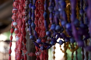 Multiple strands of handcrafted paper beads in various colors, primarily red, purple, and yellow, hang in vertical rows. The beads are textured and cylindrical in shape, with a natural and artisanal appearance. The background is softly focused, emphasizing the vibrant colors and craftsmanship of the beads.
