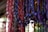 Multiple strands of handcrafted paper beads in various colors, primarily red, purple, and yellow, hang in vertical rows. The beads are textured and cylindrical in shape, with a natural and artisanal appearance. The background is softly focused, emphasizing the vibrant colors and craftsmanship of the beads.