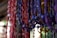 Multiple strands of handcrafted paper beads in various colors, primarily red, purple, and yellow, hang in vertical rows. The beads are textured and cylindrical in shape, with a natural and artisanal appearance. The background is softly focused, emphasizing the vibrant colors and craftsmanship of the beads.