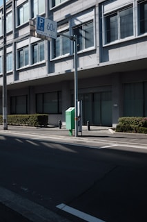 A street view featuring a modern building with large rectangular windows and a green hedge alongside the pavement. A street sign with 'P 60' in blue is mounted on a pole, along with another sign in Japanese. Shadows are cast across the street, and there is a green box with the number '60' on it beside the building.