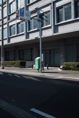 A street view featuring a modern building with large rectangular windows and a green hedge alongside the pavement. A street sign with 'P 60' in blue is mounted on a pole, along with another sign in Japanese. Shadows are cast across the street, and there is a green box with the number '60' on it beside the building.