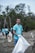 A group of people wearing blue T-shirts with the words 'Ocean Cleanup Group' engaged in a beach cleanup. They are holding large white garbage bags to collect trash. The background features trees and a cloudy sky, creating a natural environment.