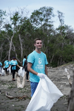 A group of people wearing blue T-shirts with the words 'Ocean Cleanup Group' engaged in a beach cleanup. They are holding large white garbage bags to collect trash. The background features trees and a cloudy sky, creating a natural environment.
