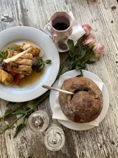 A rustic table setting featuring a plate of grilled chicken and roasted vegetables, a cup of coffee in a floral mug, and a round loaf of bread with a spoon inserted. Pink roses and salt and pepper shakers are neatly arranged beside the dishes.