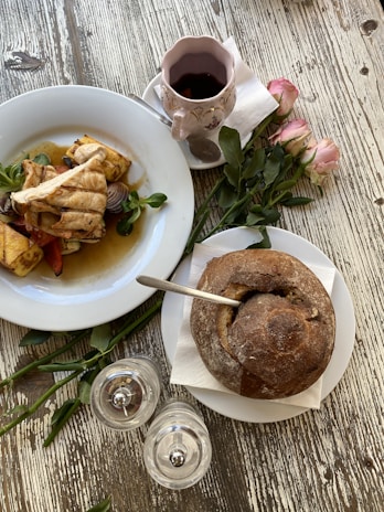 A rustic table setting featuring a plate of grilled chicken and roasted vegetables, a cup of coffee in a floral mug, and a round loaf of bread with a spoon inserted. Pink roses and salt and pepper shakers are neatly arranged beside the dishes.