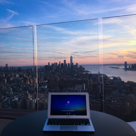A laptop sits on a table overlooking a vast cityscape during sunset. The skyline features tall skyscrapers against a backdrop of a colorful sky, blending shades of blue, pink, and orange. The scene is viewed through glass barriers, adding a reflective quality to the image.