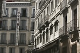 A view of old European-style buildings with intricate wrought iron balconies and a sign reading 'Hostal Persal' mounted vertically on one of the structures. The architecture is classic, with large windows and light-colored facades, suggesting a historical or traditional urban setting.