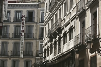 A view of old European-style buildings with intricate wrought iron balconies and a sign reading 'Hostal Persal' mounted vertically on one of the structures. The architecture is classic, with large windows and light-colored facades, suggesting a historical or traditional urban setting.