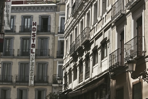 A view of old European-style buildings with intricate wrought iron balconies and a sign reading 'Hostal Persal' mounted vertically on one of the structures. The architecture is classic, with large windows and light-colored facades, suggesting a historical or traditional urban setting.