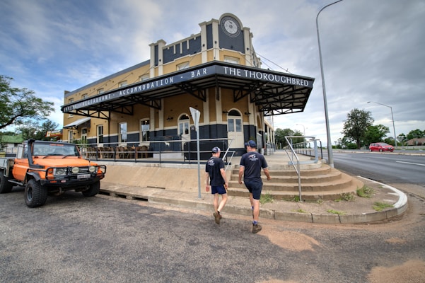 A historic-looking building with a sign reading 'The Thoroughbred' offers accommodation and dining options. Two people, casually dressed, walk toward the entrance. An orange off-road vehicle is parked on the left side of the image, while the street nearby is relatively empty except for a red car in the distance. Trees and a cloudy sky complete the scene.
