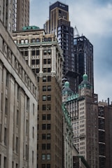 A cluster of tall, densely packed skyscrapers in varying architectural styles with intricate details and green rooftops. The scene is captured from a street-level perspective, highlighting the imposing structures against a cloudy sky.