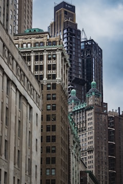 A cluster of tall, densely packed skyscrapers in varying architectural styles with intricate details and green rooftops. The scene is captured from a street-level perspective, highlighting the imposing structures against a cloudy sky.