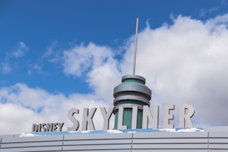 A stylized sign with the words 'Disney Skyliner' in bold, metallic letters against a backdrop of blue sky and clouds. The structure features a cylindrical, futuristic design element with a tall antenna-like spike on top.