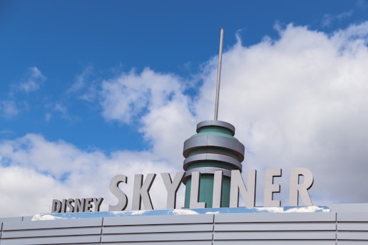 A stylized sign with the words 'Disney Skyliner' in bold, metallic letters against a backdrop of blue sky and clouds. The structure features a cylindrical, futuristic design element with a tall antenna-like spike on top.