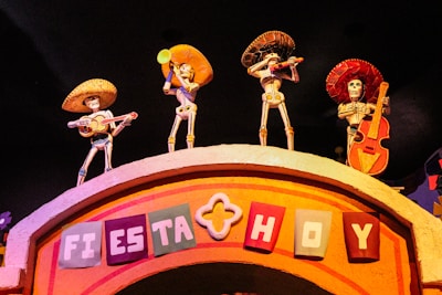 Four skeletal figures wearing sombreros and traditional Mexican attire, each playing a musical instrument, are positioned on top of an arched structure. The sign on the arch reads 'Fiesta Hoy' in colorful block letters. The setting is vibrant and festive, likely representing a cultural celebration.