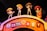 Four skeletal figures wearing sombreros and traditional Mexican attire, each playing a musical instrument, are positioned on top of an arched structure. The sign on the arch reads 'Fiesta Hoy' in colorful block letters. The setting is vibrant and festive, likely representing a cultural celebration.