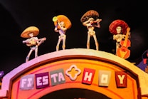 Four skeletal figures wearing sombreros and traditional Mexican attire, each playing a musical instrument, are positioned on top of an arched structure. The sign on the arch reads 'Fiesta Hoy' in colorful block letters. The setting is vibrant and festive, likely representing a cultural celebration.