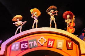Four skeletal figures wearing sombreros and traditional Mexican attire, each playing a musical instrument, are positioned on top of an arched structure. The sign on the arch reads 'Fiesta Hoy' in colorful block letters. The setting is vibrant and festive, likely representing a cultural celebration.