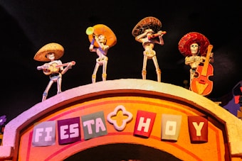 Four skeletal figures wearing sombreros and traditional Mexican attire, each playing a musical instrument, are positioned on top of an arched structure. The sign on the arch reads 'Fiesta Hoy' in colorful block letters. The setting is vibrant and festive, likely representing a cultural celebration.
