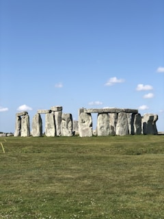 A historical site featuring a set of ancient stone structures arranged in a circle on a green grassy field, under a clear blue sky with a few scattered clouds.