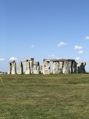 A historical site featuring a set of ancient stone structures arranged in a circle on a green grassy field, under a clear blue sky with a few scattered clouds.