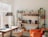 Interior of a cozy room with a workspace featuring a laptop on a wooden desk, surrounded by natural light from large windows. A comfortable chair is positioned by the desk. To the right, there is a modern shelving unit adorned with various plants, books, framed photos, and speakers. An orange armchair, draped with a blanket, adds warmth to the room.