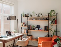 Interior of a cozy room with a workspace featuring a laptop on a wooden desk, surrounded by natural light from large windows. A comfortable chair is positioned by the desk. To the right, there is a modern shelving unit adorned with various plants, books, framed photos, and speakers. An orange armchair, draped with a blanket, adds warmth to the room.