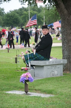 A man in traditional Scottish attire, including a kilt, is seated on a memorial bench in a park. The stone bench has a commemorative inscription and flowers are placed nearby. In the background, people are gathered, some in uniforms, and American flags are prominently displayed.