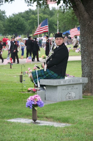 A man in traditional Scottish attire, including a kilt, is seated on a memorial bench in a park. The stone bench has a commemorative inscription and flowers are placed nearby. In the background, people are gathered, some in uniforms, and American flags are prominently displayed.