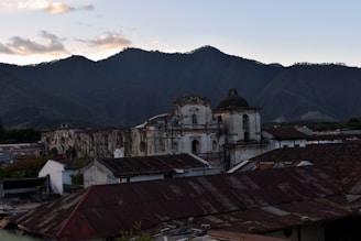 An old, weathered church stands prominently against a backdrop of lush green mountains under a clear sky. The structure features a domed roof and rustic, aged walls with visible cracks. Surrounding the church are rooftops of nearby houses, covered with red and brown tiles.