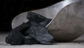 A close-up image of a metal scoop holding several pieces of black charcoal. The textured surface of the charcoal contrasts with the smooth, slightly tarnished metal surface of the scoop.