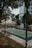 An outdoor sports court is surrounded by trees and a tall residential building. The court appears to be a mixed-use area for various sports, indicated by the painted lines. A line of benches is situated along the side of the court, and the area is cordoned off with red and white tape. Mature trees provide some shade, and the greenery adds to the urban park-like setting.