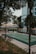 An outdoor sports court is surrounded by trees and a tall residential building. The court appears to be a mixed-use area for various sports, indicated by the painted lines. A line of benches is situated along the side of the court, and the area is cordoned off with red and white tape. Mature trees provide some shade, and the greenery adds to the urban park-like setting.