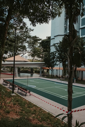 An outdoor sports court is surrounded by trees and a tall residential building. The court appears to be a mixed-use area for various sports, indicated by the painted lines. A line of benches is situated along the side of the court, and the area is cordoned off with red and white tape. Mature trees provide some shade, and the greenery adds to the urban park-like setting.