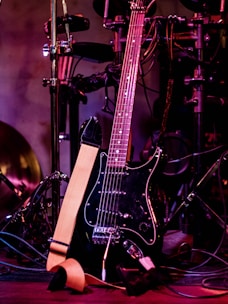A black electric guitar with a beige strap is shown resting against a set of electronic drum pads. The guitar has a glossy black finish and silver tuning pegs. The background features various musical equipment including stands and wires, with purple and red lighting adding a warm and vibrant atmosphere.