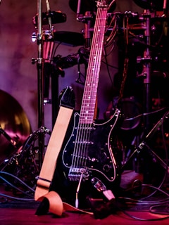 A black electric guitar with a beige strap is shown resting against a set of electronic drum pads. The guitar has a glossy black finish and silver tuning pegs. The background features various musical equipment including stands and wires, with purple and red lighting adding a warm and vibrant atmosphere.