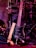 A black electric guitar with a beige strap is shown resting against a set of electronic drum pads. The guitar has a glossy black finish and silver tuning pegs. The background features various musical equipment including stands and wires, with purple and red lighting adding a warm and vibrant atmosphere.