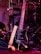 A black electric guitar with a beige strap is shown resting against a set of electronic drum pads. The guitar has a glossy black finish and silver tuning pegs. The background features various musical equipment including stands and wires, with purple and red lighting adding a warm and vibrant atmosphere.