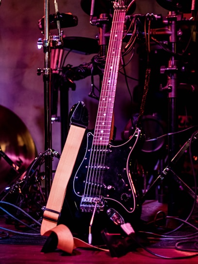 A black electric guitar with a beige strap is shown resting against a set of electronic drum pads. The guitar has a glossy black finish and silver tuning pegs. The background features various musical equipment including stands and wires, with purple and red lighting adding a warm and vibrant atmosphere.