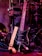 A black electric guitar with a beige strap is shown resting against a set of electronic drum pads. The guitar has a glossy black finish and silver tuning pegs. The background features various musical equipment including stands and wires, with purple and red lighting adding a warm and vibrant atmosphere.