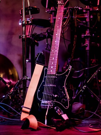 A black electric guitar with a beige strap is shown resting against a set of electronic drum pads. The guitar has a glossy black finish and silver tuning pegs. The background features various musical equipment including stands and wires, with purple and red lighting adding a warm and vibrant atmosphere.