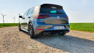 A sleek, modern hatchback car parked on a gravel road, with wind turbines visible in the background against a clear sky. The vehicle is gray with a polished finish and has sporty dual exhausts. The sun casts a bright light, emphasizing the car's curves and angles.