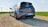 A sleek, modern hatchback car parked on a gravel road, with wind turbines visible in the background against a clear sky. The vehicle is gray with a polished finish and has sporty dual exhausts. The sun casts a bright light, emphasizing the car's curves and angles.