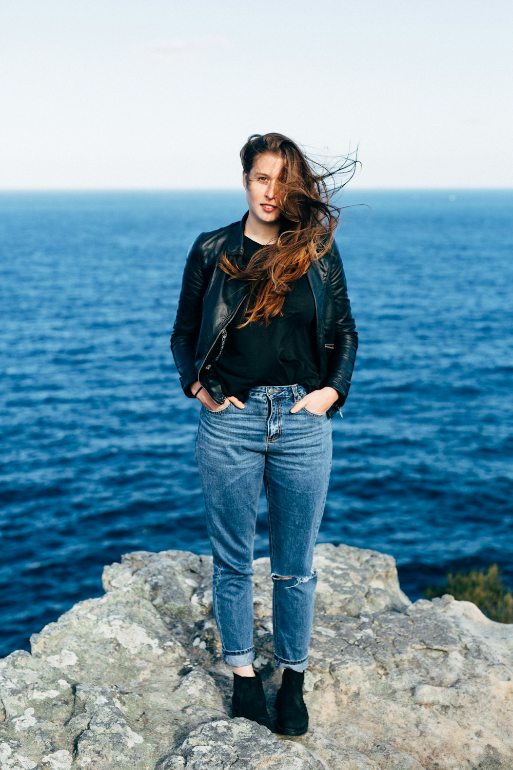 woman in black leather jacket and blue denim jeans standing on gray rock near body of near near near near