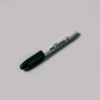 A black and white permanent marker lies diagonally on a plain, light-colored surface. The marker has a black cap and a white barrel with black text displaying the brand name.