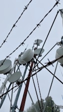 A collection of telecommunication towers with numerous satellite dishes and antennas is seen through barbed wire. The sky is overcast, creating a backdrop of gray clouds, emphasizing the industrial and slightly ominous atmosphere.