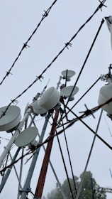 A collection of telecommunication towers with numerous satellite dishes and antennas is seen through barbed wire. The sky is overcast, creating a backdrop of gray clouds, emphasizing the industrial and slightly ominous atmosphere.
